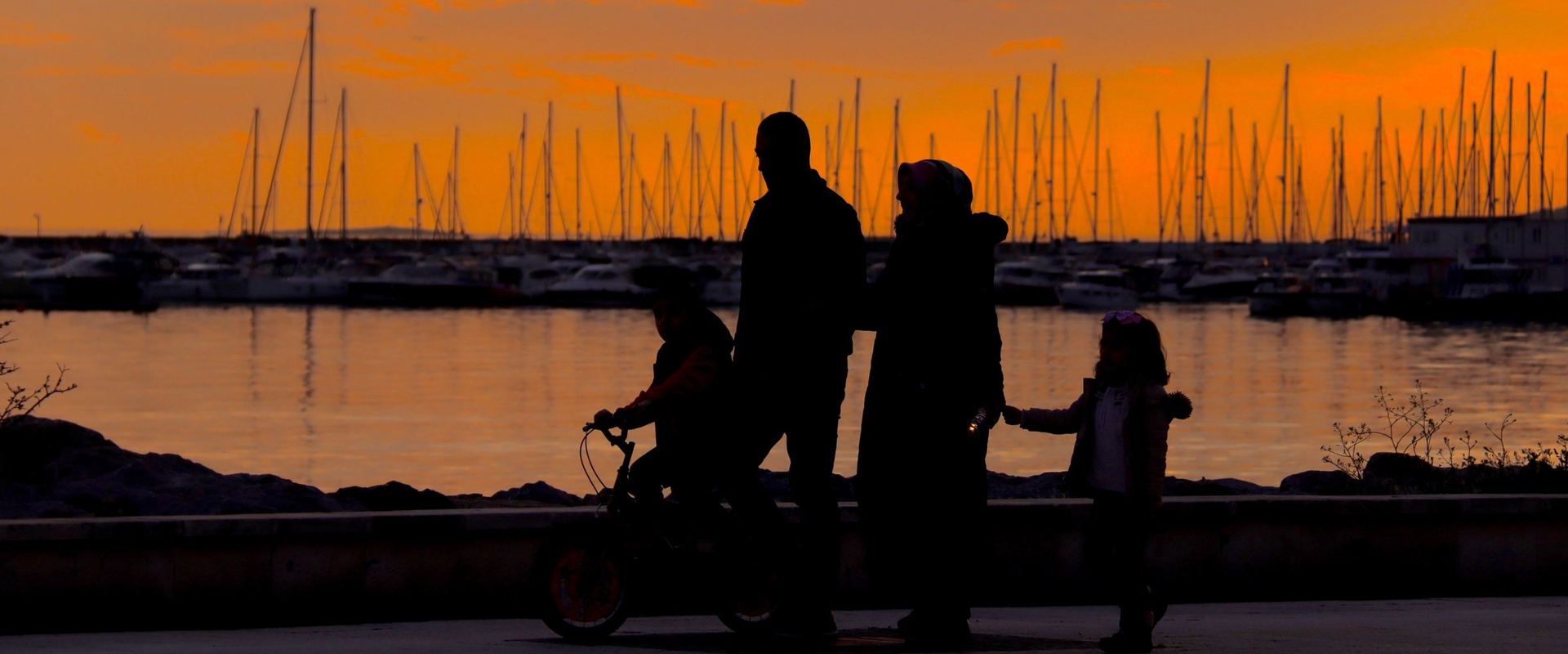 A family walking along a harbor at sunset, seen in silhouette.