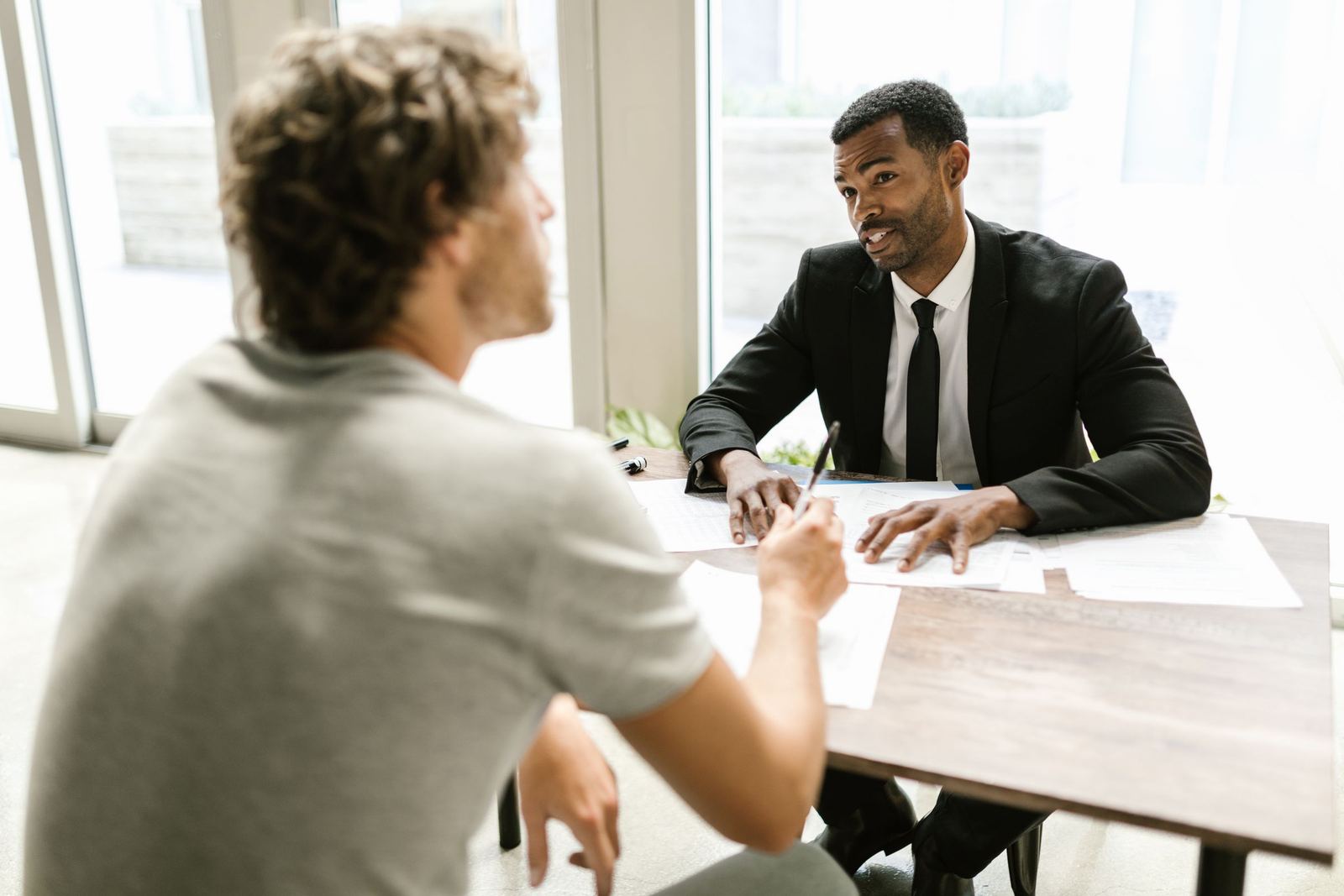 An advisor in a suit talking with a client across a desk, with paperwork between them.